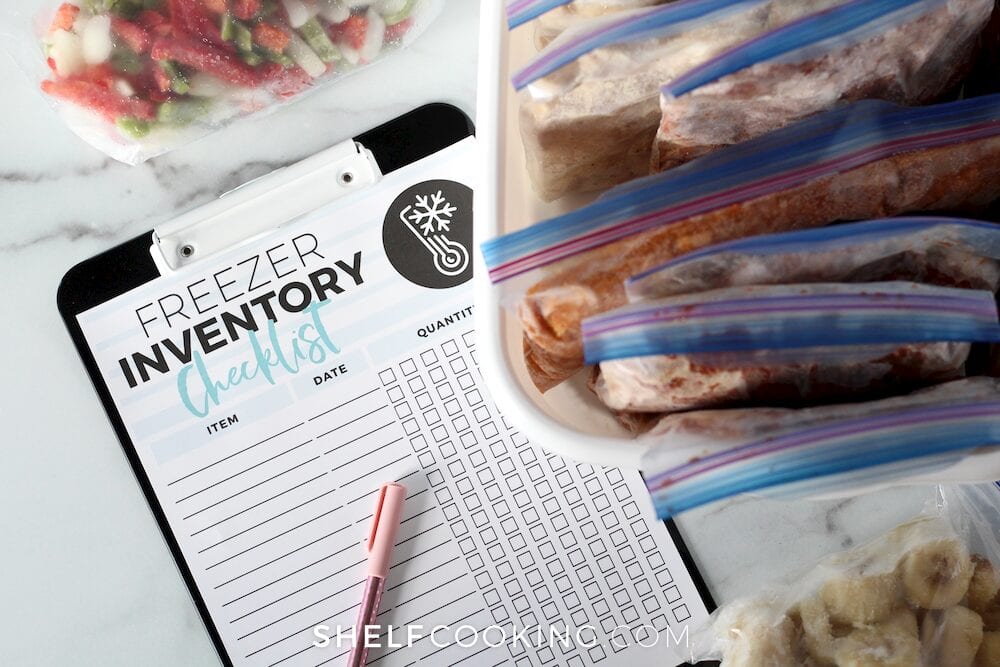 Freezer inventory checklist on a clipboard with assorted freezer bags on a counter, from Shelf Cooking 