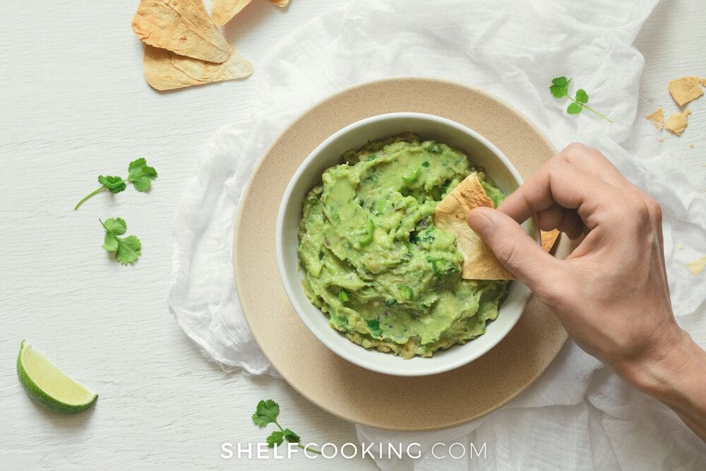 A hand dipping a tortilla chip into guacamole from Shelf Cooking