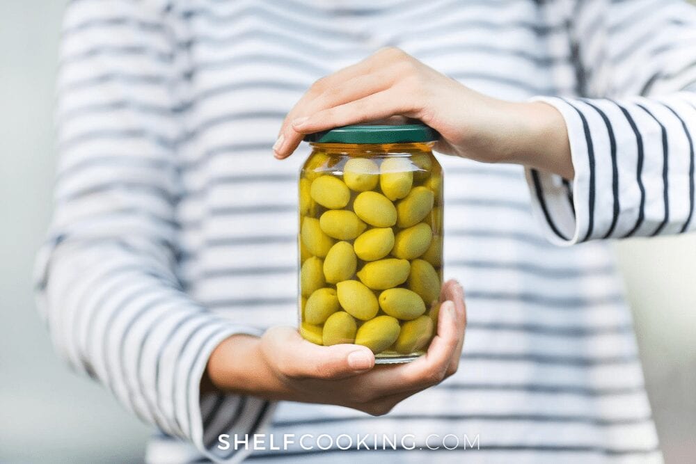 woman holding a jar of green olives with olive juice, from Shelf Cooking