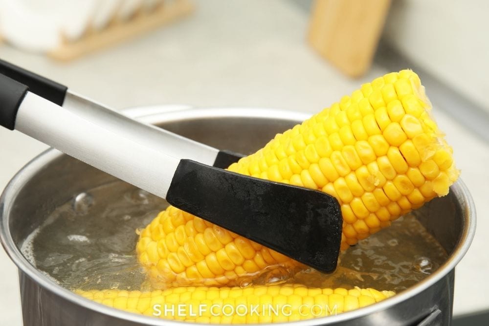 Tongs pulling corn on the cob out of a pot of boiling water from Shelf Cooking.