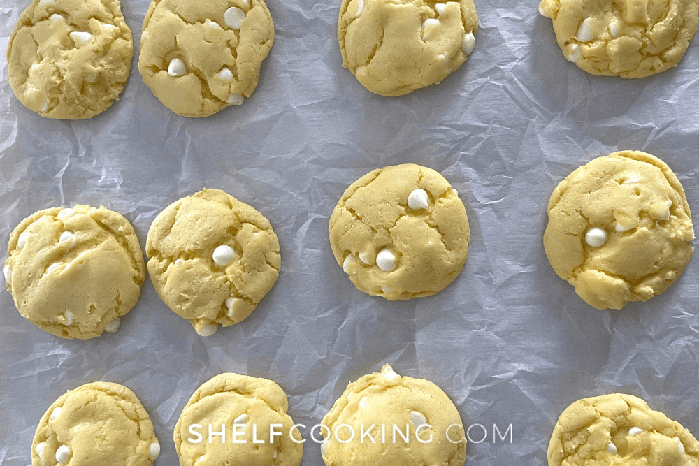 Overhead image of a dozen freshly baked lemon cake mix cookies with white chocolate chips on parchment paper on a cookie sheet.