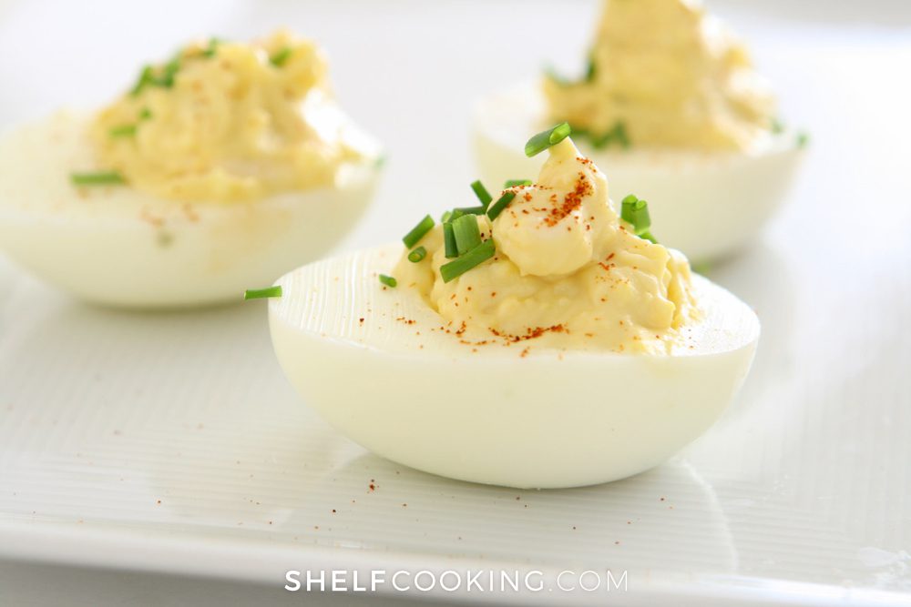 Close-up image of deviled eggs on a white platter. The yellow yolk filling is piled high with a green garnish on top. - Shelf Cooking