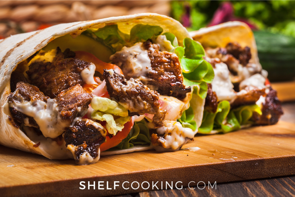 Close up of a steak burrito with lettuce, tomatoes, and sauce, on a wooden cutting board. - Shelf Cooking