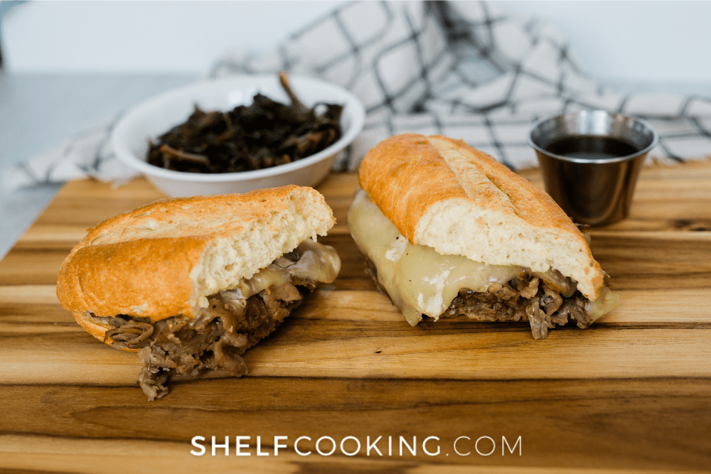 Wooden cutting board with a French dip sandwich on it, cut in half. There is a small cup of au jus and a white bowl and towel in the background. 
