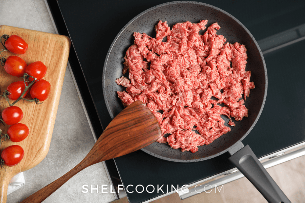 Overhead image of a frying pan on a stove with ground beef and a wooden spoon. A cutting board with red tomatoes is next to it. - Shelf Cooking