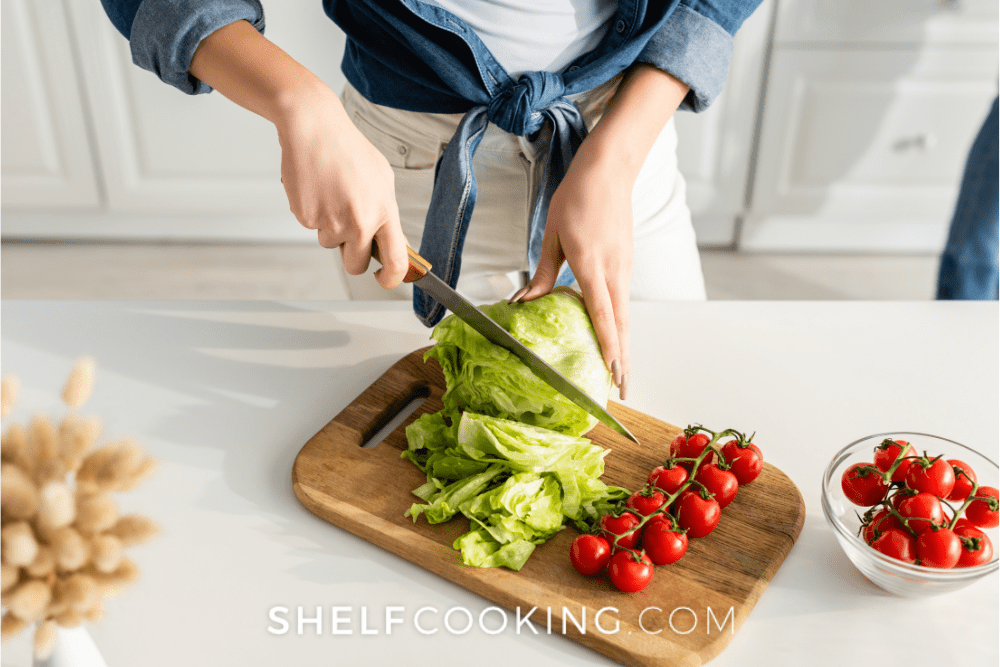 Image of woman cutting lettuce and tomatoes on a wooden cutting board. - Shelf Cooking