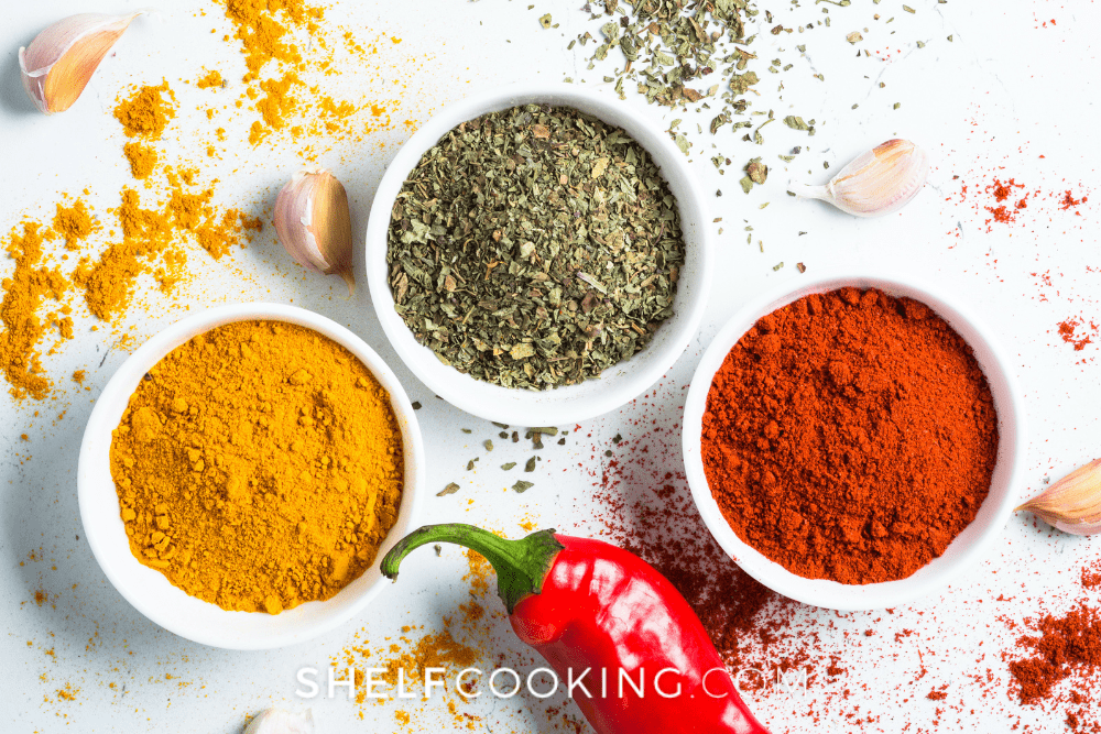 Image of three small white bowls filled with chili pepper, oregano, and cumin. Around the bowls is scattered powder, garlic cloves, and a red chili pepper. - Shelf Cooking