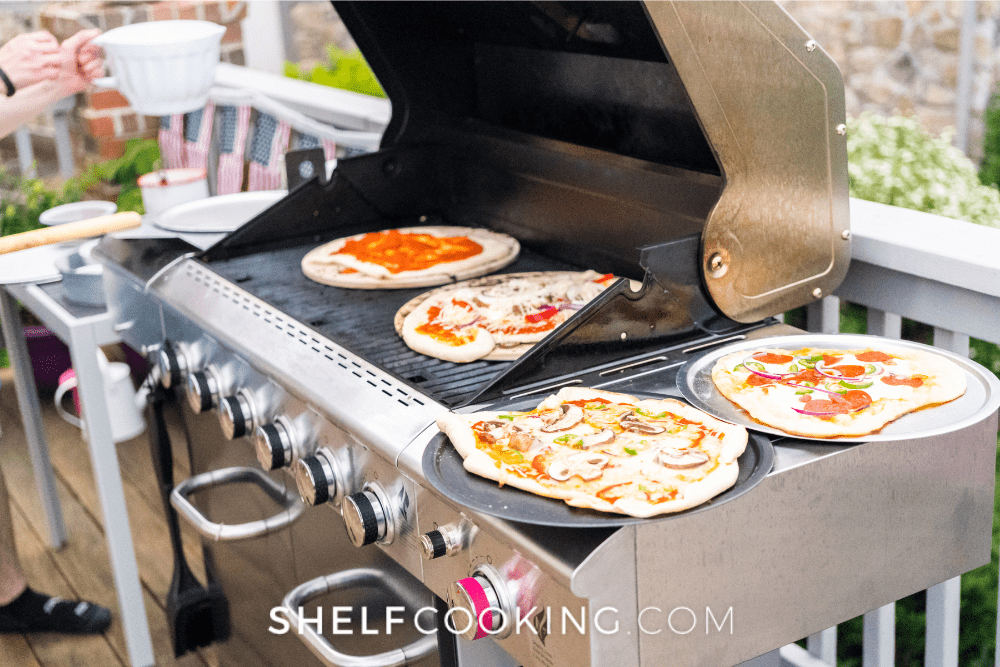 Image of large bbq grill with the lid open and pizzas placed on the grate. In the background you can see the set up for an American picnic. - Shelf Cooking