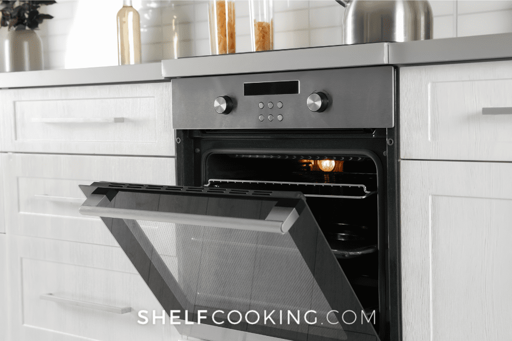 Image of a stainless steel oven with the door partially open, with white cabinets on each side and a gray countertop. - Shelf Cooking
