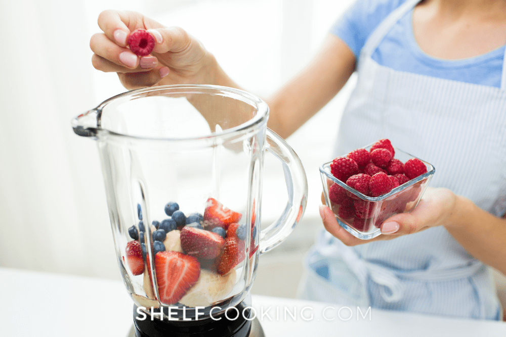 Image of girl in a blue apron filling a blender with berries and bananas. - Shelf Cooking