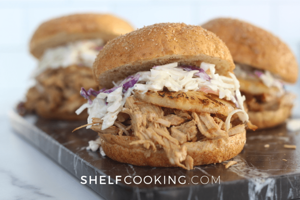 Image of a Hawaiian pulled pork sandwich with pineapple and coleslaw on top. There are two more sandwiches in the background. - Shelf Cooking