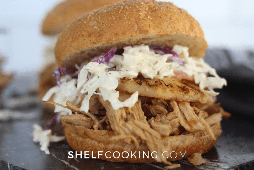 Close up image of a pulled pork sandwich topped with a grilled pineapple ring and coleslaw. 