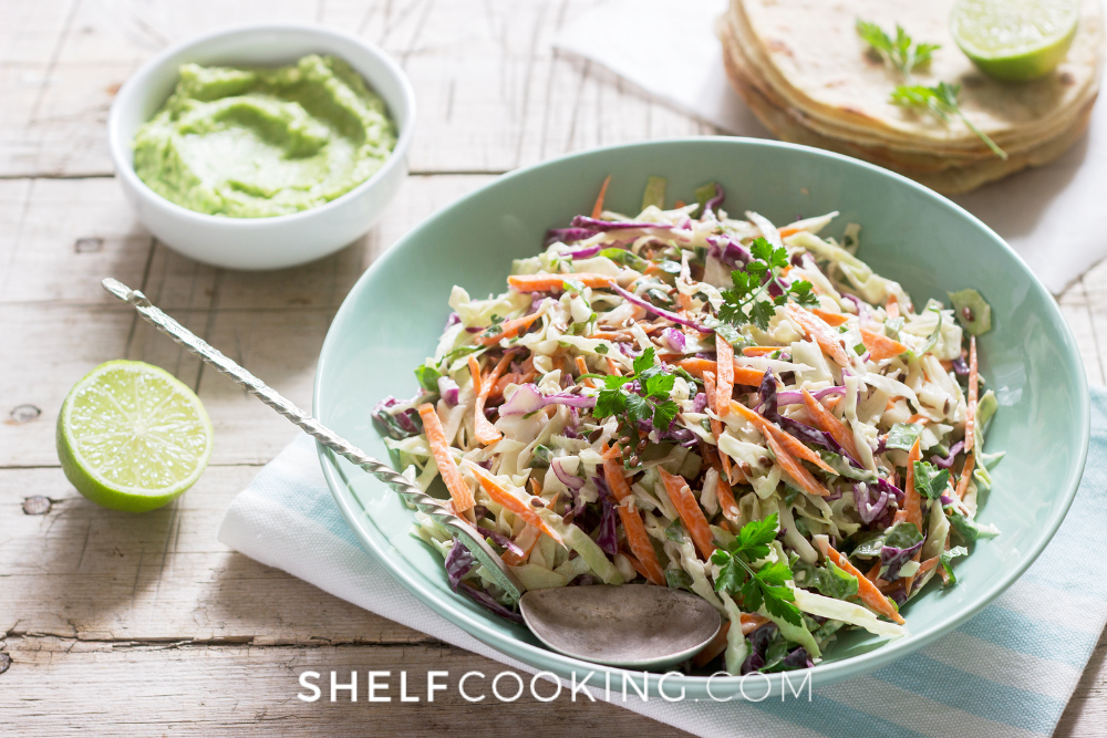 Image of a teal bowl filled with cabbage and carrot coleslaw. There is a silver spoon in the bowl, and in the background are tortillas and a bowl of guacamole next to a lime. - Shelf Cooking