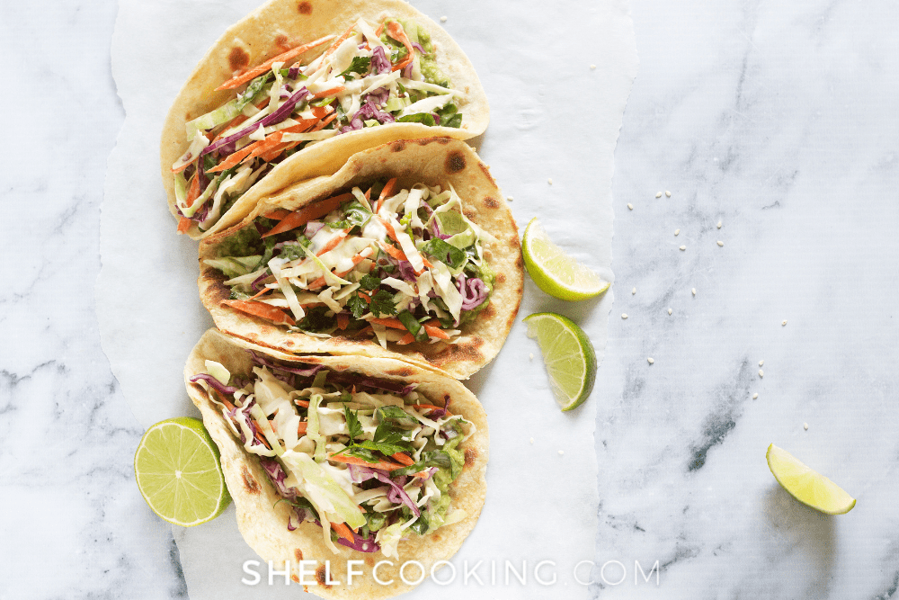 Image of three fish tacos with coleslaw on a white and grey marble countertop, sounded by lime slices. - Shelf Cooking