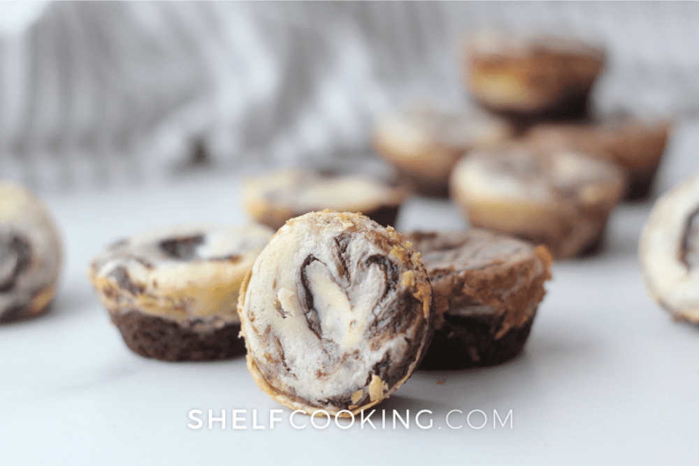 Image of several cheesecake brownie bites scattered on a white table. -Shelf Cooking