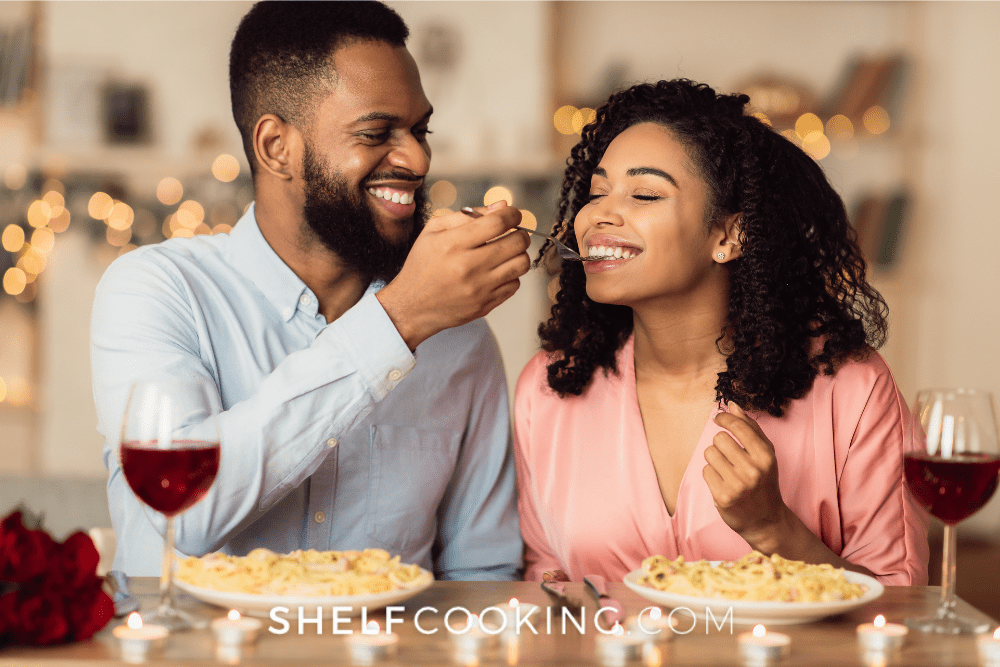 Image of a man and woman eating a pasta date night dinner at home. They are smiling as the man feeds the woman a forkful. - Shelf Cooking