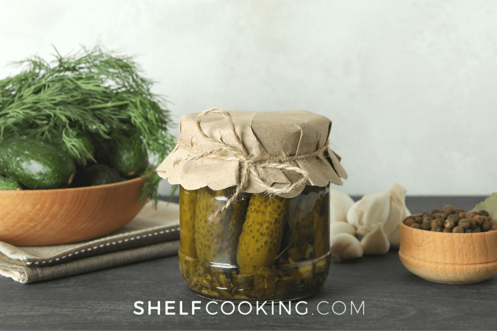 Image of a jar of homemade pickles with paper on top, held in place by twine. In the background there are spices and garlic. - Shelf Cooking