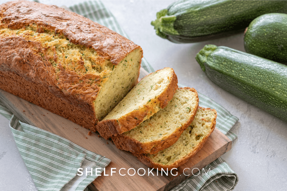 Image of a loaf of zucchini bread with a few slices cut. Zucchini squash is located to the side. - Shelf Cooking
