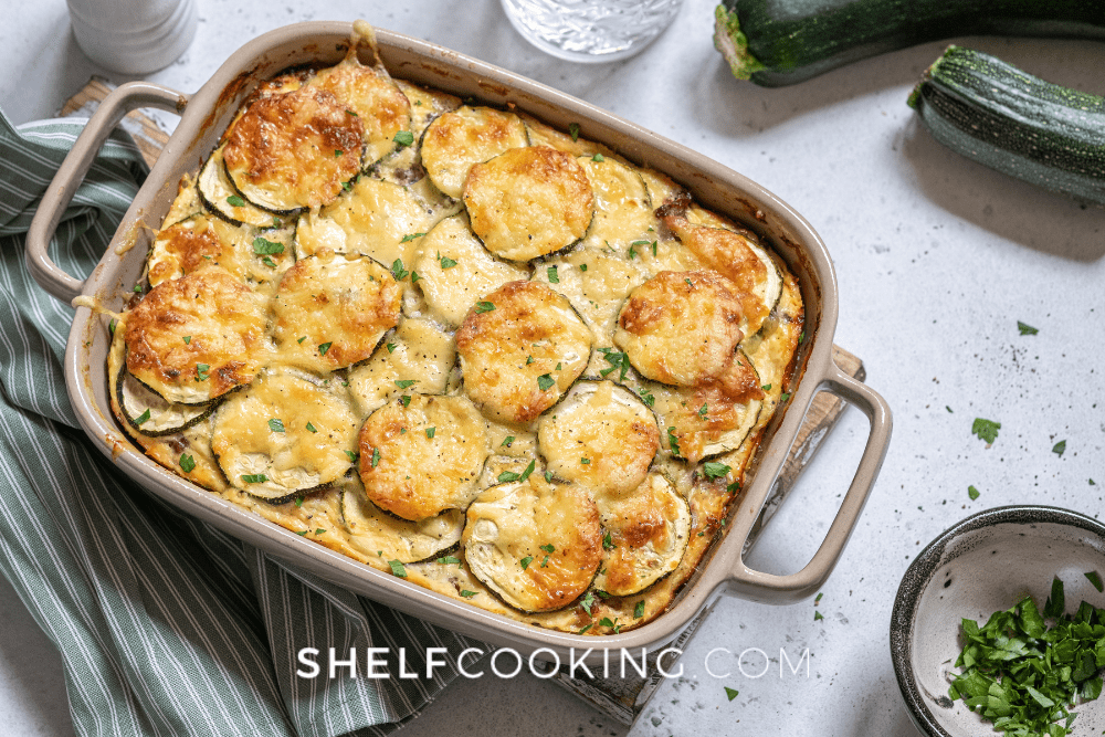Image of a casserole dish with summer squash gratin. A kitchen towel and zucchini squash are off to the side. - Shelf Cooking