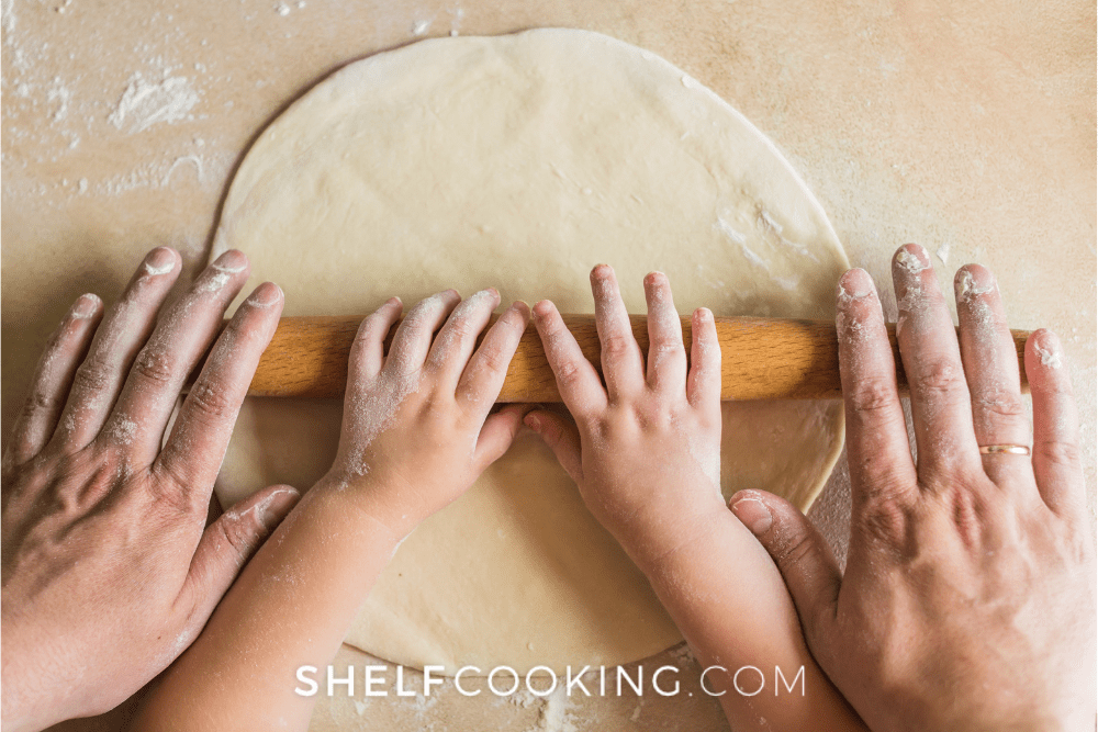 Overhead image of an adult and young child working together to roll out dough with a rolling pin. - Shelf Cooking