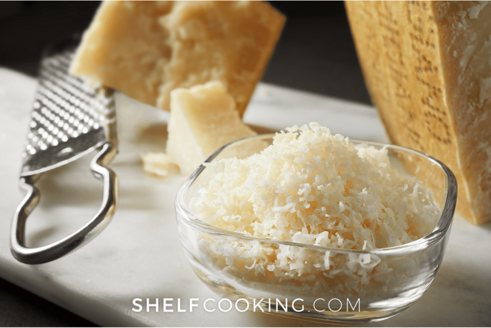 Image of glass bowl with shredded parmesan cheese. A block of parmesan cheese and a grater are in the background. - Shelf Cooking