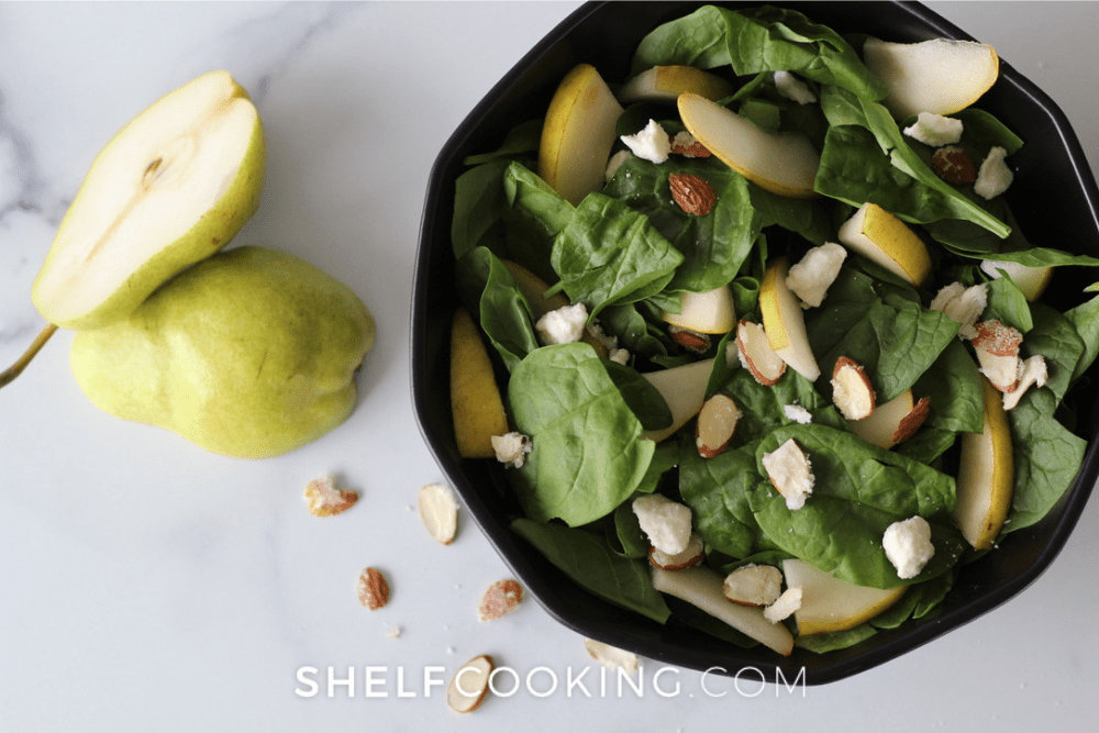 Overhead image of a fall pear salad in a bowl. A sliced pear and slivered sugared almonds are next to it. - Shelf Cooking